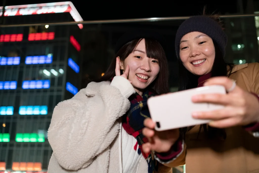 Two women taking a selfie at night with a smartphone in a city, showing social interaction and mobile-first digital behavior.
