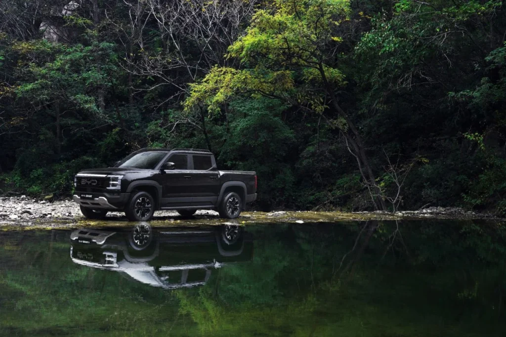Black BYD electric pickup parked beside a calm river with forest reflections