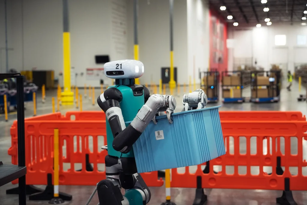 Digit humanoid robot transporting a blue storage tote through an active warehouse environment.