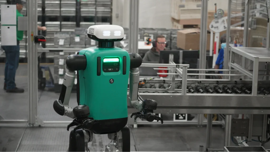 Agility Robotics' Digit robot navigating a manufacturing facility alongside human workers and conveyor systems.