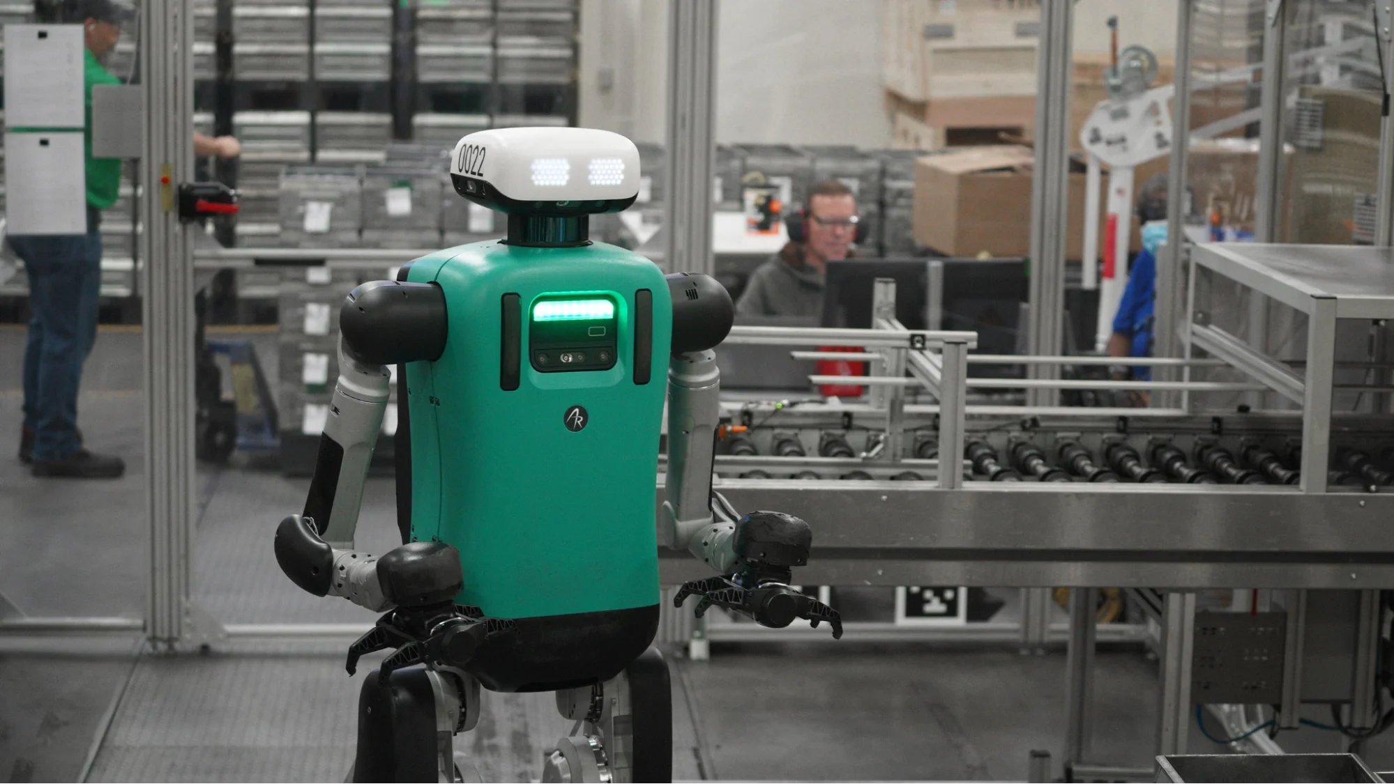 Agility Robotics' Digit robot navigating a manufacturing facility alongside human workers and conveyor systems.