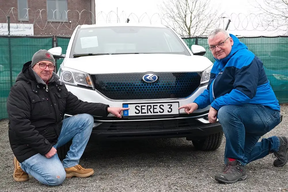 Two men posing beside a white SERES 3 SUV pointing at the front badge