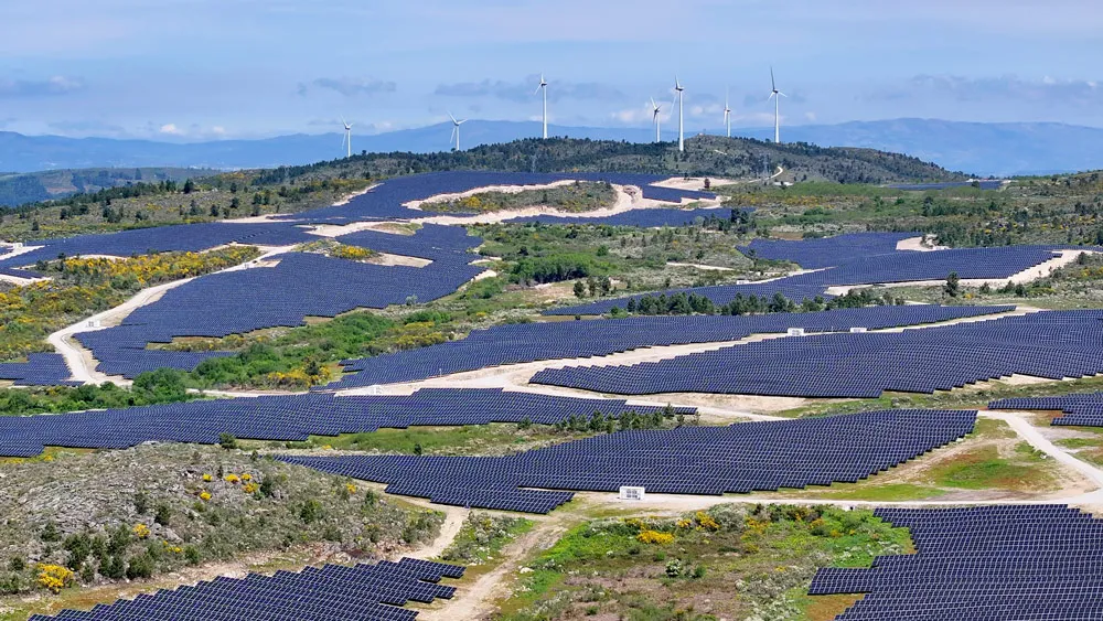 Hillside renewable energy site with large solar arrays and wind turbines in the background.