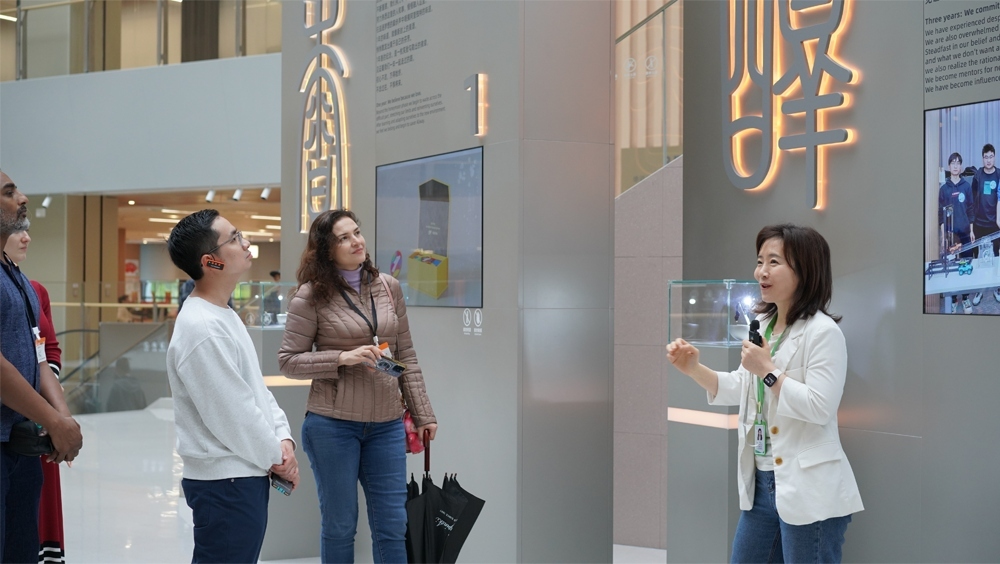 A guide holding a microphone speaks with visitors beside exhibition displays.